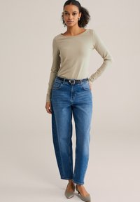 Šviesiai smėlio spalvos ilgomis rankovėmis palaidinė, derinama su šviesiai mėlynais, tiesių kojų džinsais, turinčiais priekinės kišenes ir juodą diržą su sidabrine sagtimi.