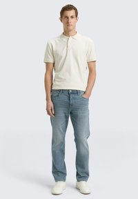 Koszulka polo w jasnobeżowym kolorze z teksturowanym wykończeniem, zestawiona z jasnoniebieskimi dżinsami i białymi trampkami. Prosty, klasyczny swobodny strój.