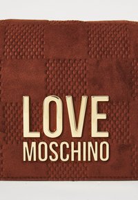 Braune strukturierte Lederoberfläche mit goldmetallicfarbener "LOVE MOSCHINO"-Buchstabierung, die in der Mitte platziert ist.