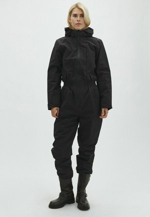 Femme aux cheveux blonds portant une combinaison à capuche noire et des bottes noires, debout devant un fond clair uni.