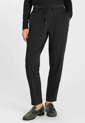 Persona che indossa pantaloni neri a taglio affusolato con coulisse in vita e mocassini neri in pelle, in piedi su uno sfondo bianco uniforme.