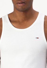 Hvit bomullstopp med rund hals og en liten brodert logo i rødt, hvitt og blått på brystet.