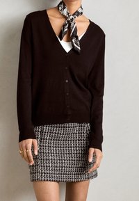 Svart cardigan med v-ringad hals, knappframsidan och långa ärmar, tillsammans med en mönstrad svartvit tweedkortkjol. Silkscarf knuten runt nacken.