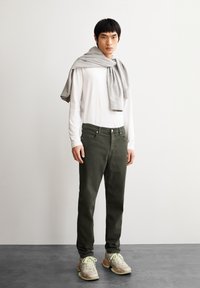 Khaki broek met voorzakken, gecombineerd met een witte langemouwen shirt. Een lichtgrijs vest over de schouders gedrapeerd. Sneakers met multicolor accenten.