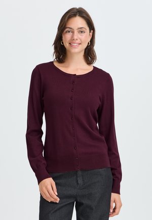 Femme aux cheveux bruns mi-longs porte des boucles d’oreilles créoles dorées, un cardigan violet foncé boutonné et un pantalon gris foncé, debout devant un fond blanc.