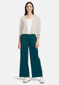 Cardigan en crème texturé, chemise blanche et pantalon large teal. Modèle portant des baskets blanches. Tissu doux avec des bords effilochés sur le cardigan.