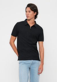 Melna polo krekls ar rāvējslēdzēja kaklu, īsām piedurknēm un pieguļošu dizainu. Valkāts ar gaiši ziliem džinsiem uz neitrāla fona.