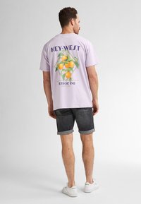 Camiseta morada de manga corta con un gráfico colorido de cítricos en la parte posterior, combinada con pantalones cortos de mezclilla oscuros enrollados y zapatillas blancas.