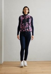 Steppweste mit floralem Muster in den Farben Pink und Lila, schwarzer Hintergrund, kombiniert mit dunklen Jeans und weißen Sneakers.