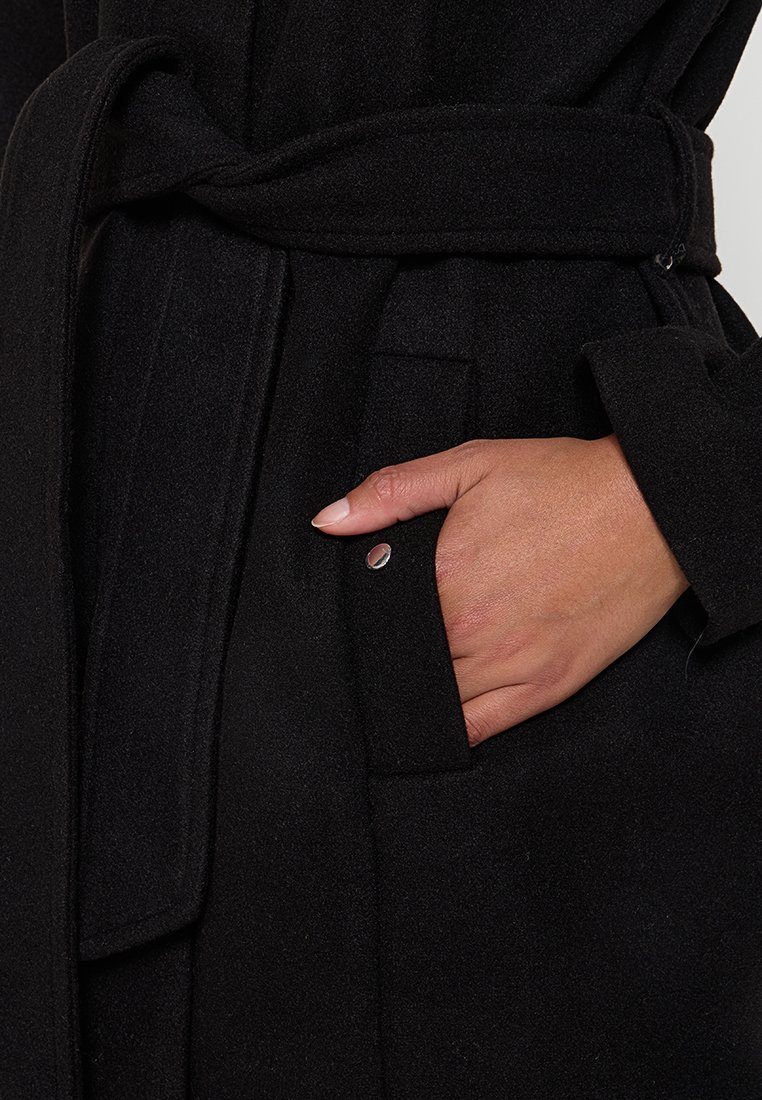 Un manteau noir en tissu doux et texturé, avec une ceinture nouée et une poche latérale avec un accent en argent. Accent sur un design utilitaire et pratique.