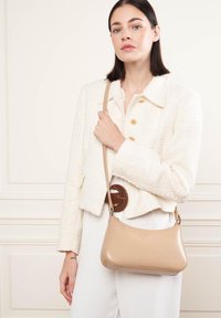 Sac à épaule beige avec une texture lisse, une forme arrondie et une longue bandoulière. Le sac est porté avec une tenue blanche comprenant une veste en crème texturée.