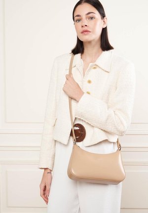 Sac à épaule beige avec une texture lisse, une forme arrondie et une longue bandoulière. Le sac est porté avec une tenue blanche comprenant une veste en crème texturée.
