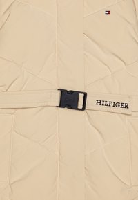 Beige gewatteerde jas met een gestikte ontwerpelement, een ceintuur in de taille met een zwarte gesp, en HILFIGER branding in zwarte stiksels.