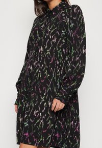 Czarna sukienka z długim rękawem w kolorowy, abstrakcyjny wzór w odcieniach zieleni, różu i fioletu. Posiada kołnierzyk i projekt z falbaniastym dołem.