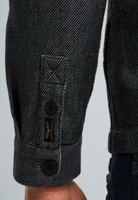 Polsino di camicia in denim nero con motivo a zigzag, due bottoni neri e una piccola etichetta con logo con dettagli di cucitura testurizzati.