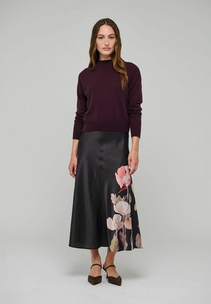Kobieta ubrana w ciemnofioletowy sweter, czarną satynową spódnicę z różowym kwiatowym nadrukiem i brązowe spiczaste buty, stojąca na tle jednolitego jasnego tła.