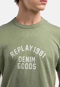 Camiseta de algodón verde oliva con cuello redondo; presenta texto impreso en blanco: "REPLAY 1981 DENIM GOODS" en disposición de arco.
