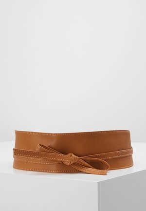 Ceinture de taille en cuir marron avec un corps large et une fine lanière nouée en nœud sur une surface d'exposition blanche.
