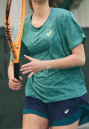 Personne portant une chemise verte et un short bleu marine tenant une raquette de tennis orange, se préparant à frapper une balle de tennis en intérieur.
