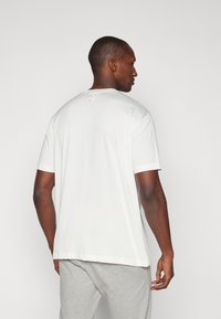 Camiseta blanca de algodón con mangas cortas, cuello redondo y un pequeño detalle decorativo en la parte posterior del cuello. Textura suave, corte relajado.