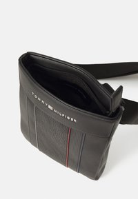 Black leather crossbody bag with zip closure, textured front, red and blue accent stripes, and logo on the front panel. Adjustable strap included.