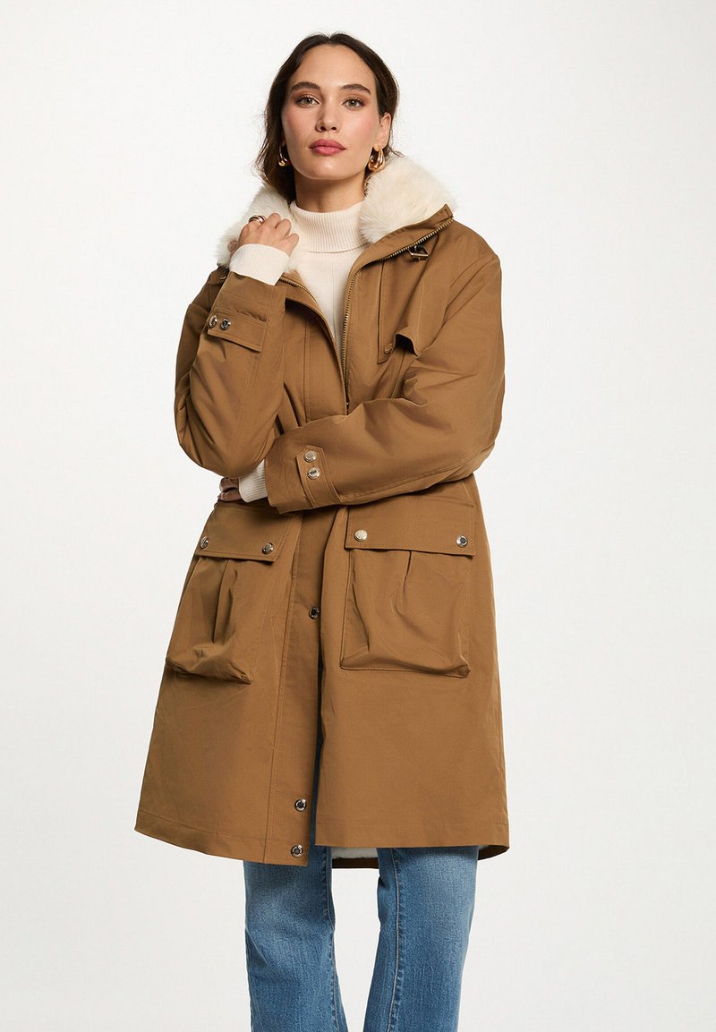 Parka marrone con colletto in pelliccia sintetica, chiusure con zip e bottoni a pressione, grandi tasche frontali e vestibilità rilassata. Indossato sopra un dolcevita color crema.