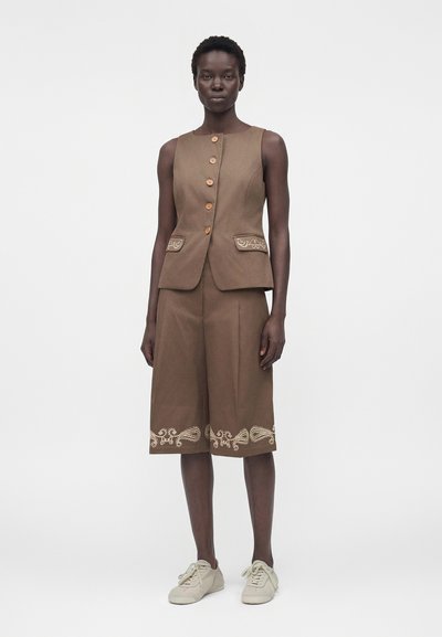 Γυναίκα που φορά αμάνικο καφέ γιλέκο με κουμπιά και ασορτί σορτς μέχρι το γόνατο με ελαφριά κεντητά σχέδια, συνδυασμένα με μπεζ αθλητικά παπούτσια.