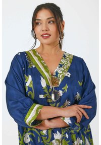 Blauwe bloemen tuniek met v-hals, groene en witte patroondetails, en losse mouwen. Lichte stof met witte bloemen en verfijnde ontwerpen.