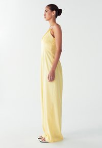 Mujer con el cabello recogido en un moño, usando pendientes de aro dorados y un vestido amarillo largo sin mangas, de pie de perfil sobre un fondo blanco.