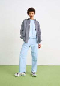 Sweat-shirt bleu clair et pantalon assorti, superposés avec une chemise rayée marine et blanche. Baskets avec des accents gris et noir. Sol vert.
