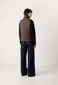 Kvinne med kort hår iført en brun quiltet vest, svart kabelstrikket genser og vide mørkeblå jeans som står mot en hvit bakgrunn.