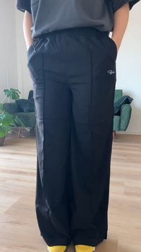 Osoba v nosení voľných čiernych nohavíc a žltých tenisiek stojí v obývacej izbe so zelenými kreslami a izbovou rastlinou v kvetináči.