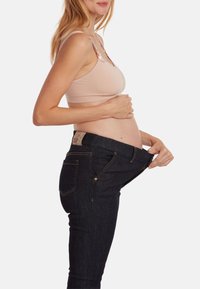 Jeans de maternité, denim foncé avec des coutures visibles, portés lâchement avec la taille baissée. Le modèle porte un soutien-gorge de couleur claire.