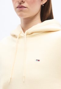 Κοντινό πλάνο ενός ατόμου που φοράει κρεμ φούτερ με κουκούλα, με ένα μικρό ορθογώνιο λογότυπο στο στήθος και ένα σκουλαρίκι κρίκο στο αριστερό αυτί.