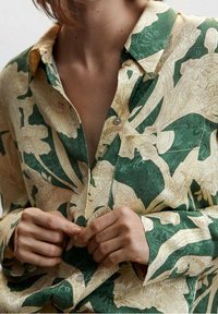 Chemise à boutons florale avec une base crème et des motifs verts, présentant un col classique et un tissu lisse, avec des coutures et des boutons visibles.