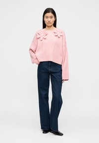 Ροζ πλεκτό πουλόβερ με τρισδιάστατες λεπτομέρειες λουλουδιών στους ώμους, συνδυασμένο με σκούρα μπλε παντελόνια φαρδιάς γραμμής και μαύρα παπούτσια.