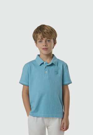 Jeune garçon aux cheveux châtain clair courts portant un polo bleu clair et un pantalon blanc, debout devant un fond uni gris clair.