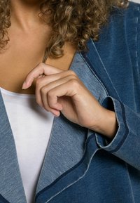 Giacca di jeans blu con una texture morbida, caratterizzata da un colletto a scialle e cuciture blu a contrasto. Mano che afferra delicatamente i capelli.