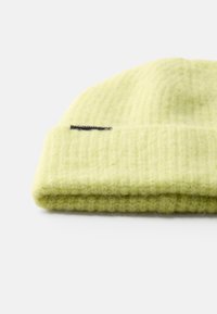 Căciulă pliată de culoare verde lime deschis, realizată dintr-un material tricotat moale și texturat. Prezintă un eticheta neagră cusută pe margine.
