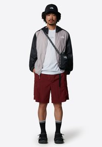 Lightweight, grey and black zip-up jacket, white t-shirt, maroon cargo shorts, black shoes with socks. Accessories include a crossbody bag and cap.