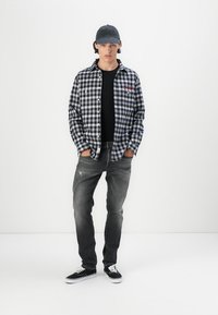 Geruite flanellen overhemd in marineblauw en wit, zwart t-shirt, grijze spijkerbroek met een lichte slijtage, en zwarte sneakers. Grijze pet met logo.