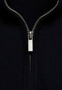 Mornarska zip-up jopica v bližnjem planu, ki ima kovinski zadrgo in teksturirano pletenino. Zadrga ima srebrno obleko in kontrastno šivanje.