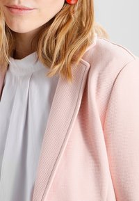 Žena s blond vlasy po ramena, oblečená v červených náušnicích, bílé blůze a strukturovaném světle růžovém blejzru na šedém pozadí.