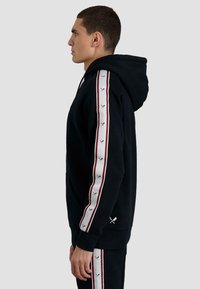 Sudadera con capucha negra con un bolsillo frontal, con rayas laterales blancas y burdeos decoradas con un patrón de espadas cruzadas en la manga. Tejido suave.