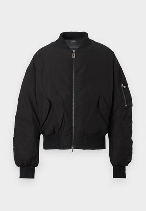 Man draagt een zwarte oversized bomberjack, losse zwarte broek, stevige zwarte schoenen, zonnebril, staand tegen een effen witte achtergrond.