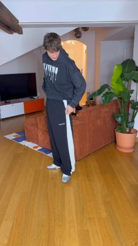 Sorte joggebukser med grå side stripe, parret med en matchende sort hættetrøje. Sølv sneakers, indendørs miljø, trægulv, og en plante synlig.