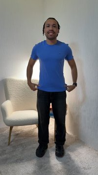 Man in blue t-shirt and black pants stands indoors, pointing at his shirt, next to a white upholstered armchair and a carpeted floor.
