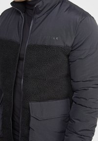 Svart vadderad jacka med en texturerad svart fleecepanel, dragkedjestängning och sidofickor. Har en slät överdel med logotypdetalj.