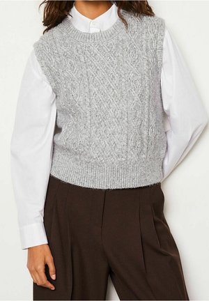 Femme portant un pull sans manches en maille torsadée gris clair sur une chemise blanche boutonnée et un pantalon taille haute marron foncé.