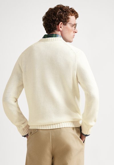 Κρεμ πλεκτό πουλόβερ με ριμπ λαιμόκοψη και μανσέτες, με άνετη εφαρμογή. Φορεμένο πάνω από ένα πουκάμισο με πράσινο καρό μοτίβο και μπεζ παντελόνι.